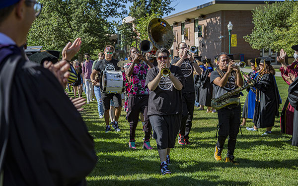 a marching band in jeans and t-shirts plays tuba, trumpets, saxophones, and base drums marches across the Quad while faculty in regalia cheer on the sidelines