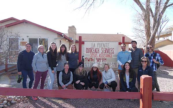 a group of 14 students and organizers smile for the camera while standing in front of a sign reading "La Puente Outreach Services Crisis Assistance"