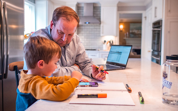 A man and a small boy sit at a kitchen counter and look at a phone surrounded by markers, paper, and a laptop