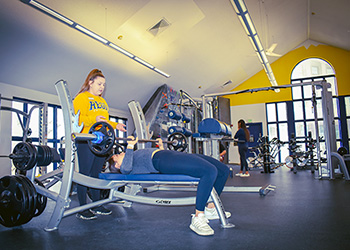 a personal trainer spots a client laying on a weight bench as he lifts a large bar with weights  in the RegisCARES clinic on the Northwest Denver campus