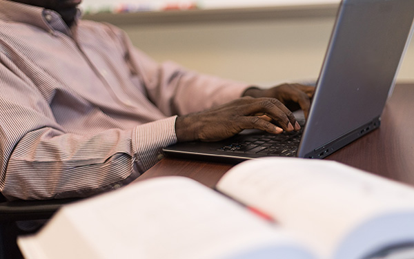 a man types on a laptop beside an open book in a classroom