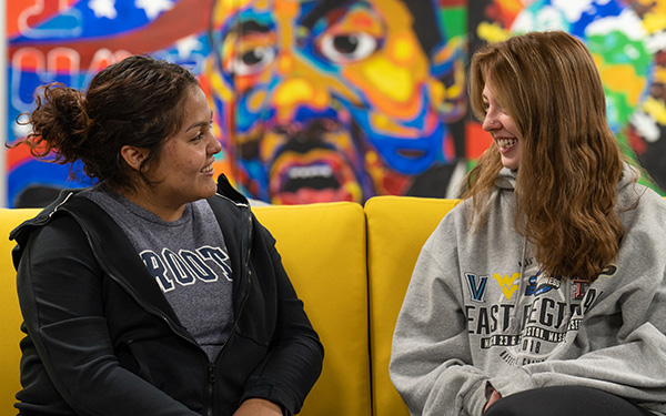 Two students chat and smile on a sofa with a colorful mural of Martin Luther King, Jr. in the background