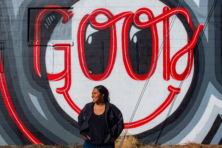 woman stands in front of a colorful mural that reads "GOOD"