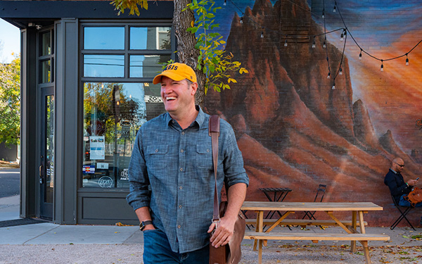 adult professional in casual clothes wears a Regis ballcap and carries a leather messenger bad while leaving a coffee shop