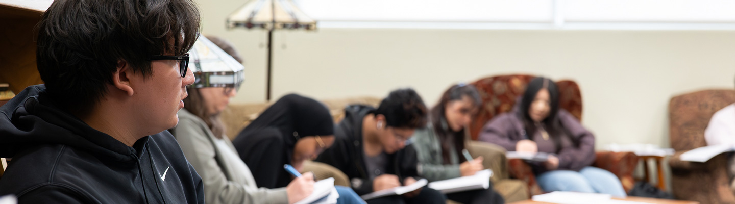 Students in creative writing sit in a row while focusing on their notebook and pen. Frontmost student looks at the front of the classroom