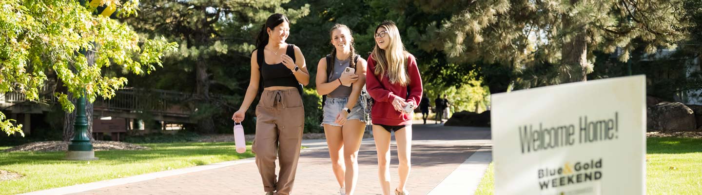 Three students walking on the Regis Campus. Yard sign that readys, Welcome Home! Regis University