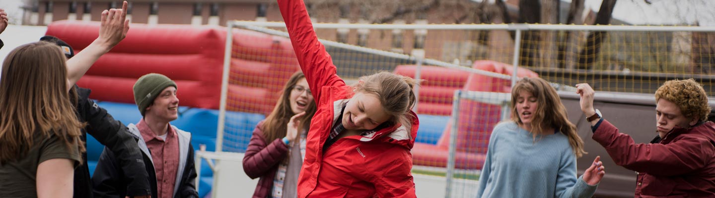 Students dance and celebrate outdoors near an inflatable structure on a cloudy day, wearing warm clothing and enjoying the event.