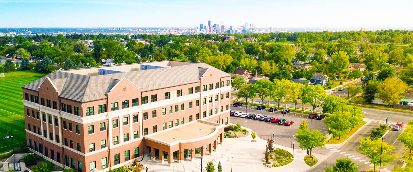 Aerial view of a modern brick academic building on a tree-lined campus, with the Denver skyline visible in the background.