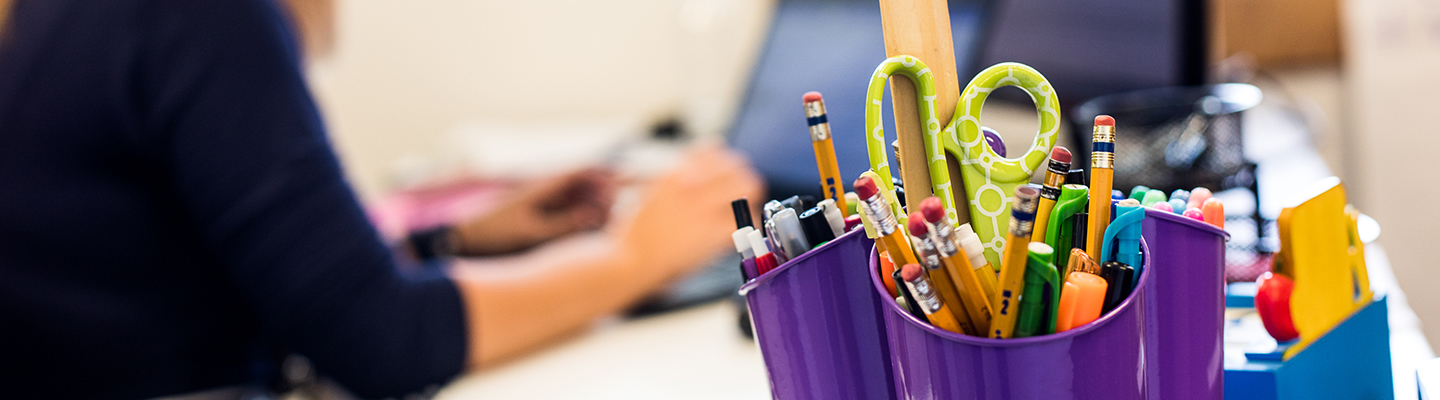 scissors and pens in holder, teacher on computer behind