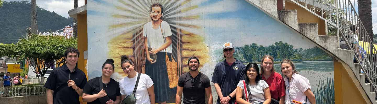 A group of Regis students stand smiling in front of a mural depicting a radiant figure with beams of light