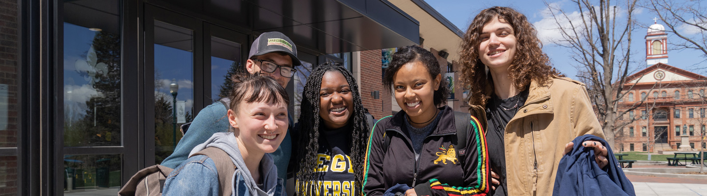 Five students stand together outside a campus building at Regis University, smiling on a sunny day with Main Hall in the background.
