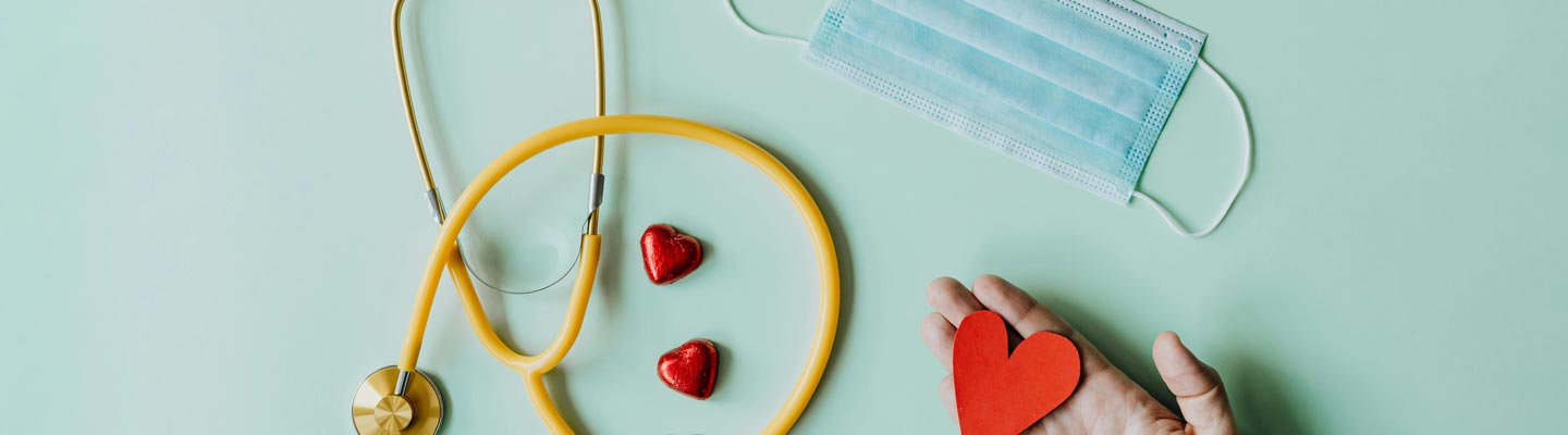 Stethoscope on the left with two candy hearts next to it. Face mask in the top right and a hand holding a paper heart at the bottom