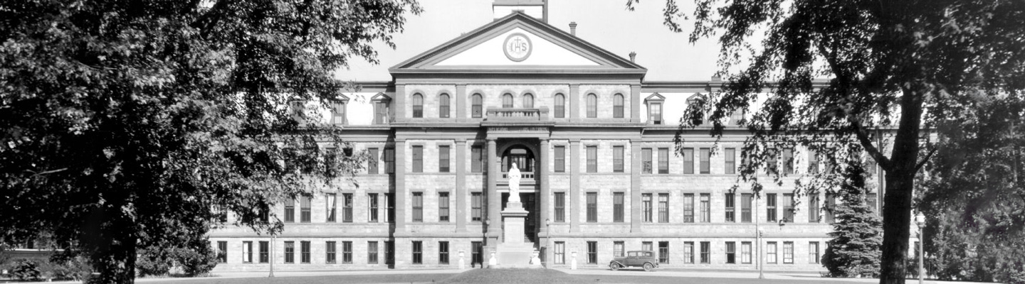 Historic black and white photo of Main Hall with trees and greenery surrounding and a old-model vehicle parked in front