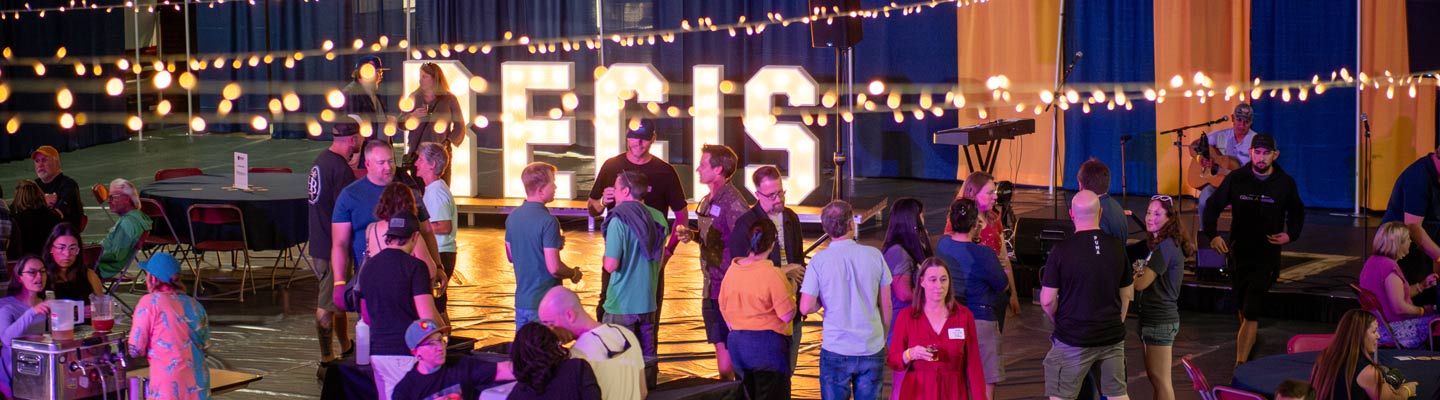 A wide, panoramic view of an indoor evening event at Regis University, featuring groups of people mingling under strands of warm string lights, with large, glowing REGIS marquee letters on a stage in the background.