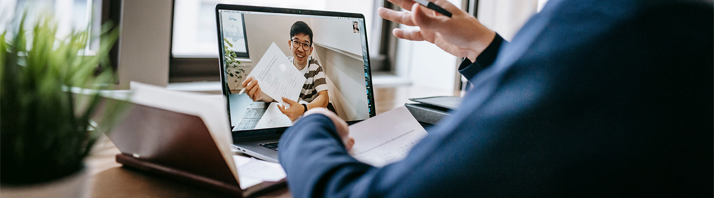 a student in an online degree program is displayed on a laptop screen holding up a class assignment in an online session with the professor
