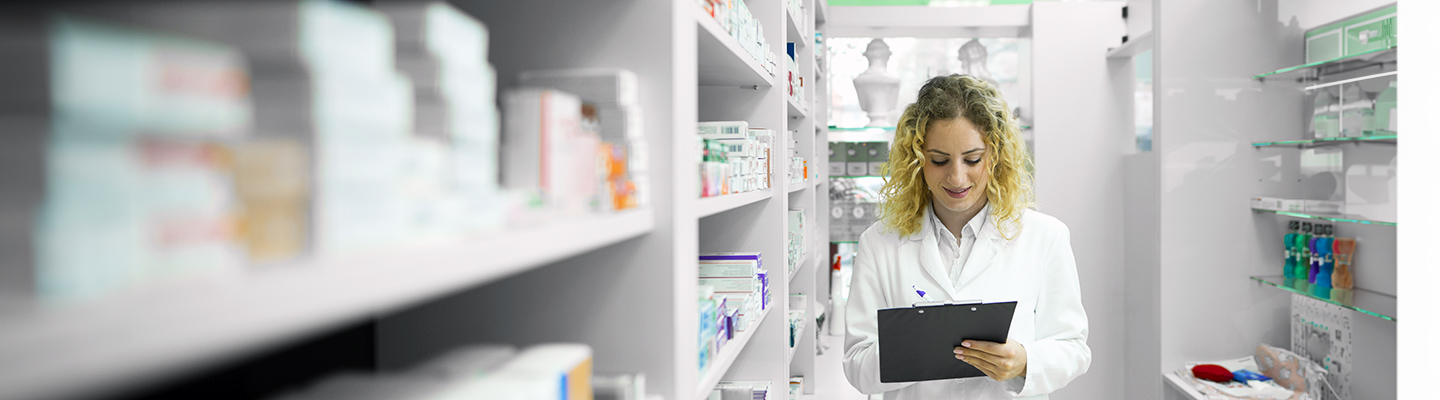 woman wearing a white lab coat and writing on a clipboard while standing in a pharmacy surrounded by shelves of medications