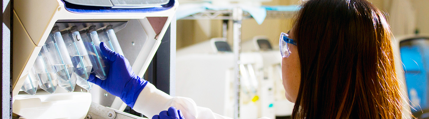 a pharmacy technician in a white lab coat, gloves and safety glasses reaches into a piece of lab machinery