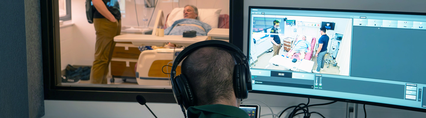 A person in a control room monitors a physical therapy simulation through a window and on a screen. A patient in a hospital bed is attended by a Regis student