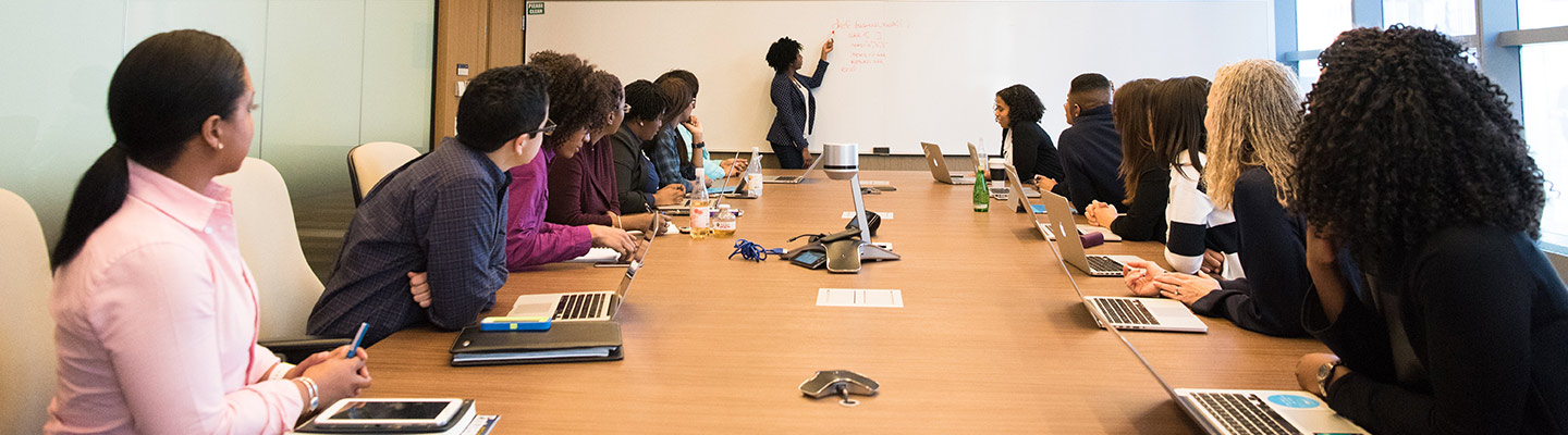 professionals seated around a large conference table listen to presenter writing on whiteboard