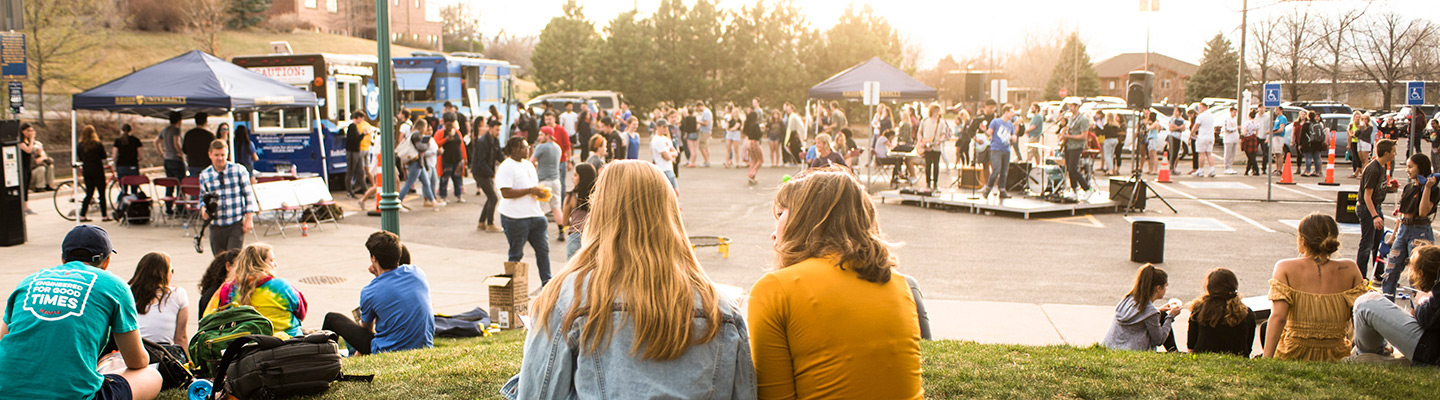 On a sunny day, a large crowd gathers in a parking lot on the Regis campus enjoying food trucks and vendors while a band sets up for a show