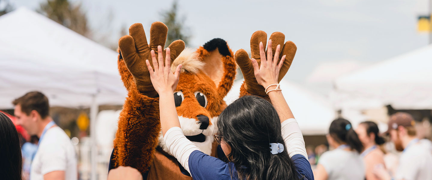 a student gives Regis fox double high fives outdoors during Welcome Week