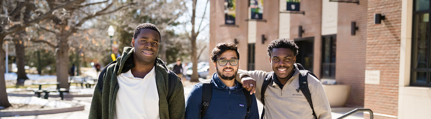 three students smile for the camera while standing outside the Student Center on the Northwest Denver Campus