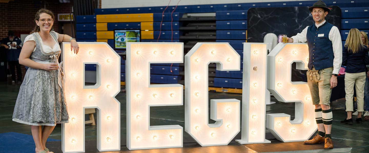 Two people dressed in traditional Bavarian-style outfits pose next to large illuminated letters spelling REGIS at Regis University's IggyBeerFest event held in a gymnasium
