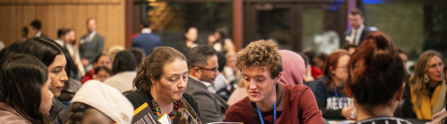 Parent and student sit at a table reading booklet while surrounded by people at their table and other tables in Mountain View Room