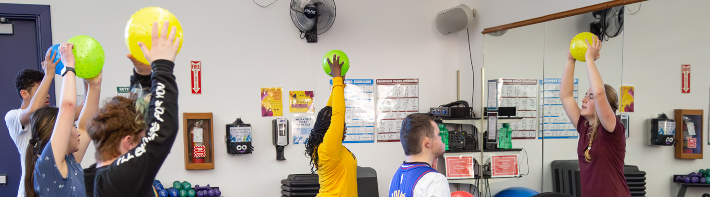 A group of people in a gym class holds colorful exercise balls above their heads