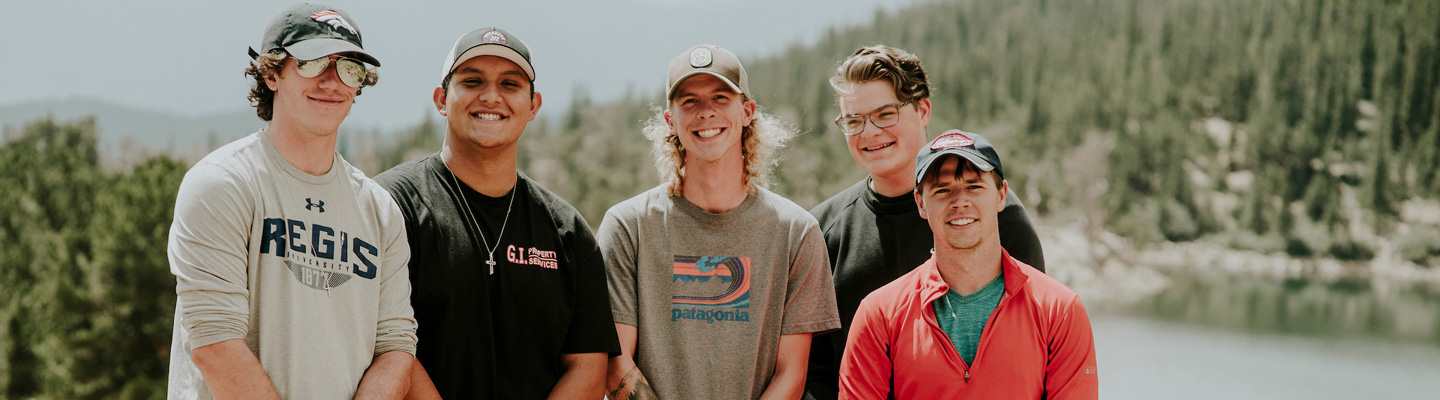 Five smiling young men posing together outdoors near a mountain lake surrounded by pine trees, several wearing baseball caps and casual clothing including a Regis University shirt.