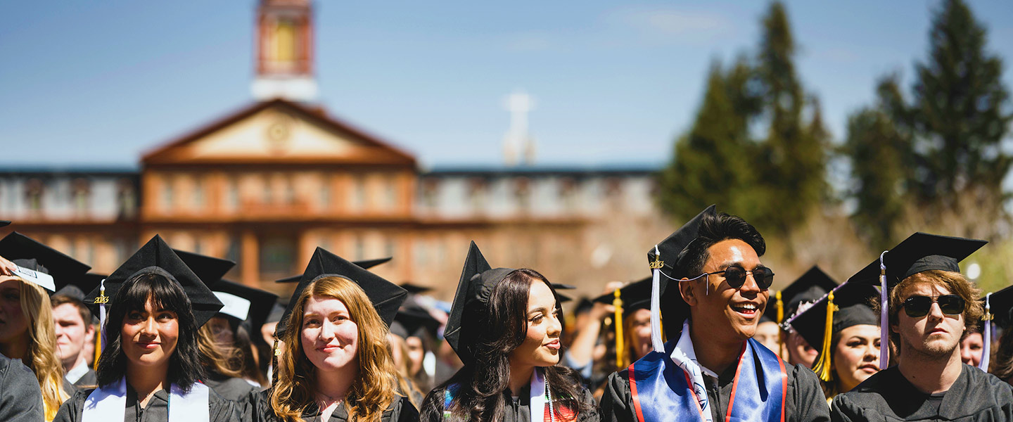 a line of students in their graduation robes and caps smiles with Main Hall in the background at commencement on the Quad