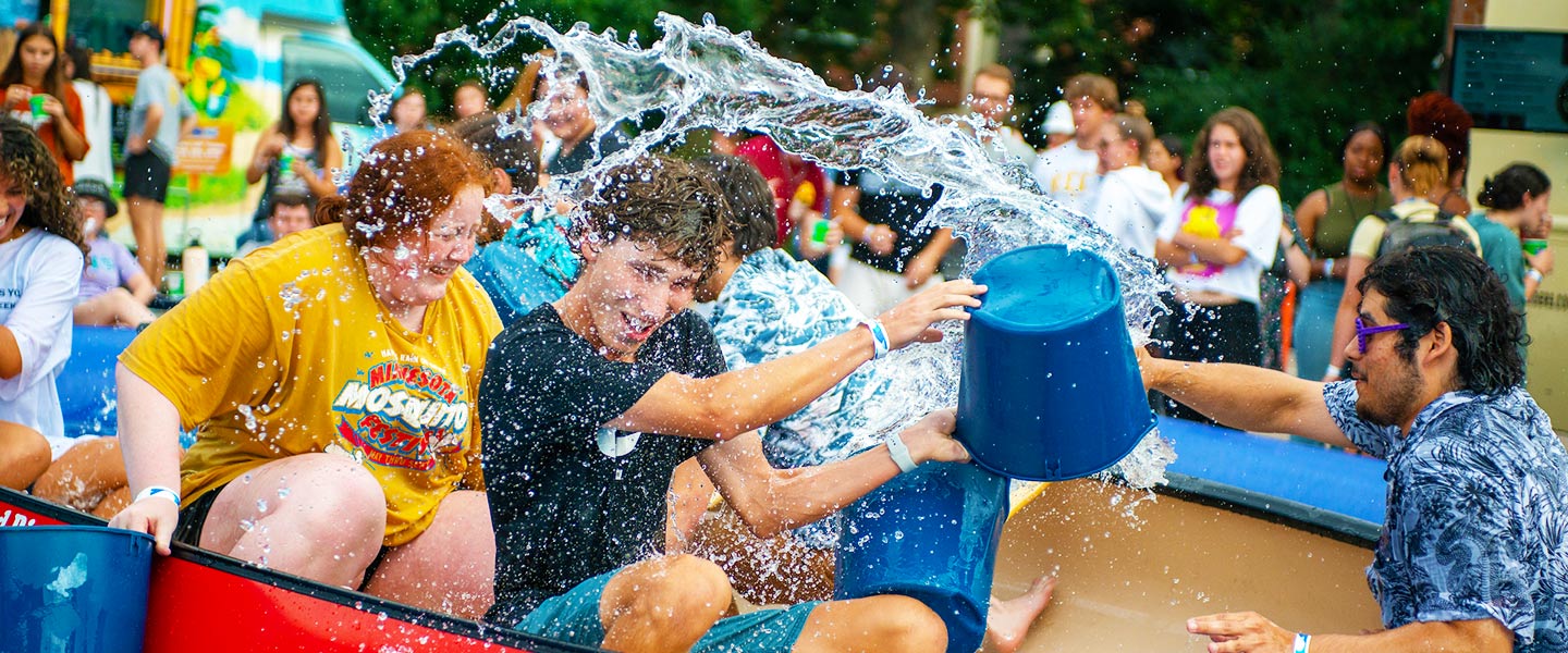 students floating in a boat on a large pool splash water on each other during Welcome Week games on campus