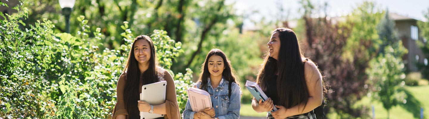 Three students walk together on the Regis campus holding school supplies with trees and bushes around them