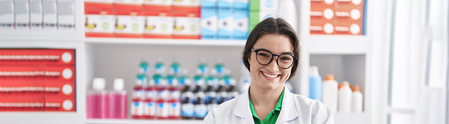 a young pharmacist wearing a white coat smiles while standing in a pharmacy with medications on the shelves
