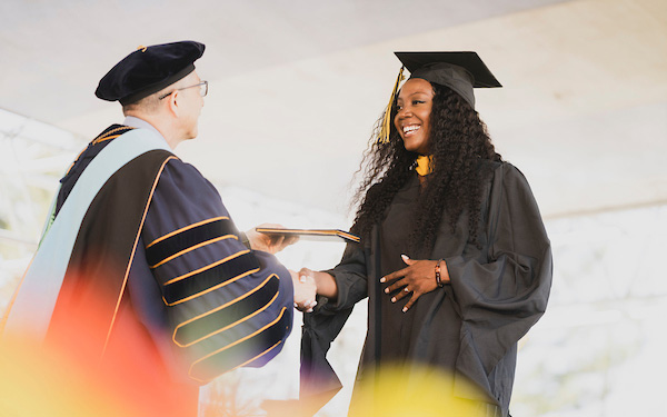 a woman wearing cap and gown accepts her diploma from the President of the University on the stage at the commencement ceremony