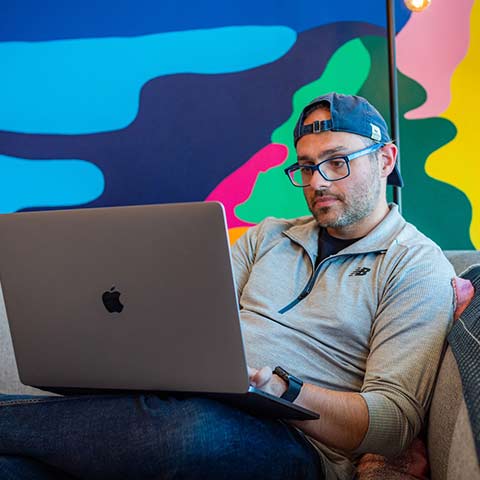 man works on a laptop while seated on a sofa in front of a colorful mural