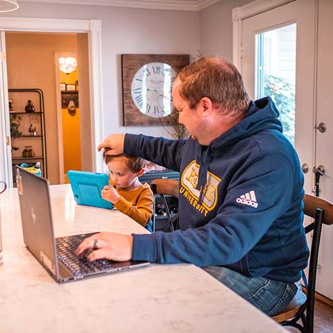 a young man wearing a Regis sweatshirt sits at the kitchen counter of his home working on a laptop. A young child sits beside him, looking at a tablet while the man ruffles his hair.
