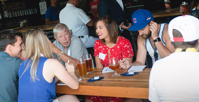 a group of men and women talk and smile while seated at a picnic table covered with glasses at a brewery