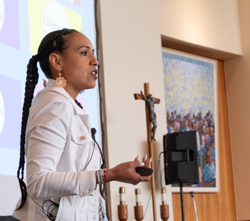 Speaker in a white jacket presents at the AJCU Diversity and Equity Network Conference, standing in front of a large screen with colorful slides.