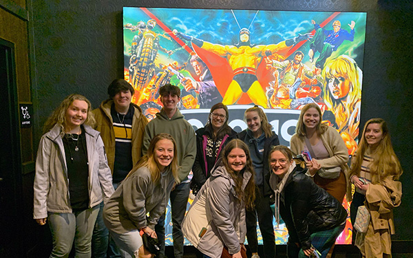 a smiling group of 9 students and professor Alyse Knorr post in front of a movie poster during their SOAR excursion to see Wakanda Forever