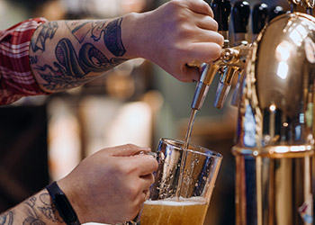 a man with tattooed arms fills a glass using a tap at a brewery