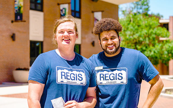 two Admissions ambassadors wearing Regis t-shirts smile on a sunny day on Boettcher Commons on campus