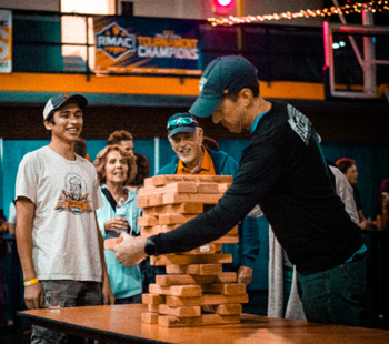 A group of people watches intently as a man in a black long-sleeve shirt and baseball cap carefully removes a wooden block from a large, unsteady Jenga tower at an indoor event.