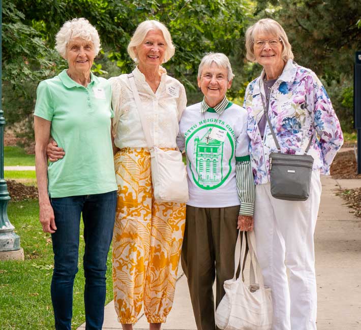 Four smiling elderly alumnae standing arm-in-arm on a campus sidewalk during a Regis University reunion.