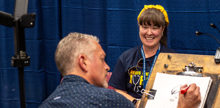 An artist draws a live caricature or portrait of a smiling woman wearing a Regis University nursing shirt and a bright yellow headband against a blue curtain backdrop.