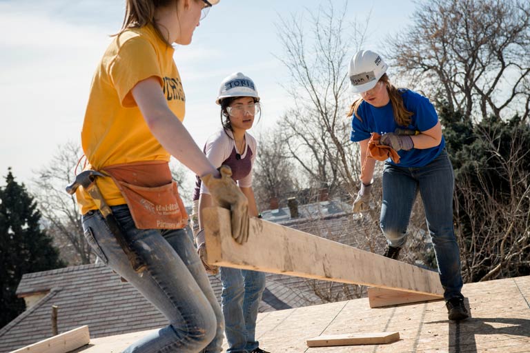 Students in hard hats carrying lumber while volunteering on a construction build.