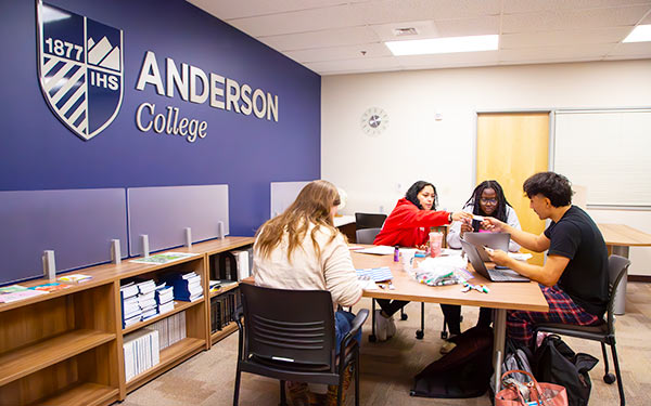 four students sit at a table collaborating on a group project in a classroom with the Anderson College of Business logo on the wall