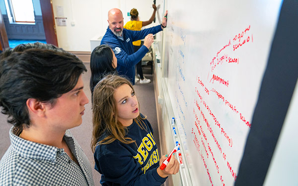 students and their professor work in pairs to brainstorm on a whiteboard in a business classroom on the Regis campus