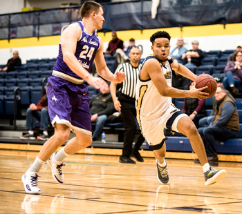 Regis University men’s basketball player in white dribbles past an opposing player in purple during a game in the campus gym.