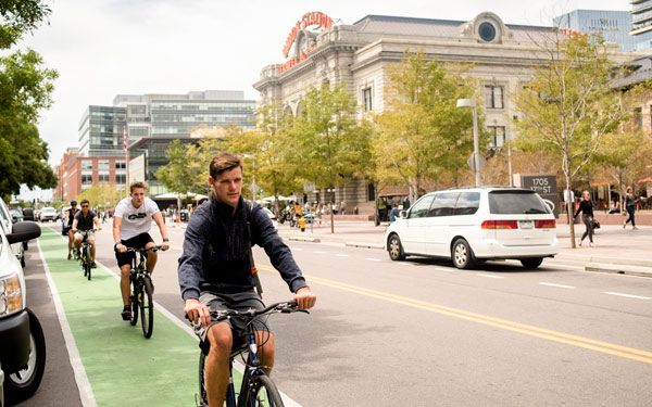 Students ride bikes in a designated green bike lane near Denver's Union Station, surrounded by city buildings and pedestrians.